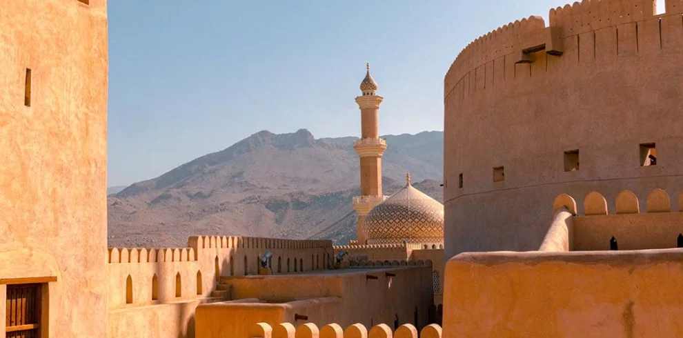 The walls and round tower of Nizwa Fort, with a mosque and mountains behind