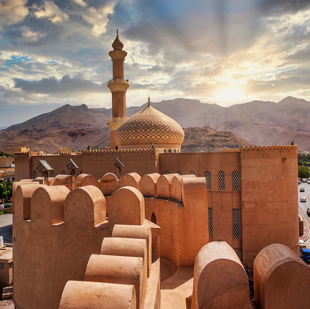 Mud-brick walls of Nizwa Fort at sunset, with a mosque dome and minaret behind