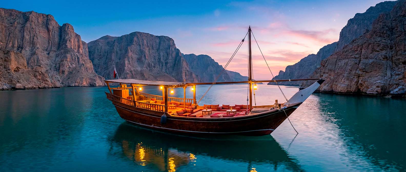 A dhow lit up at dusk in a fjord between steep cliffs
