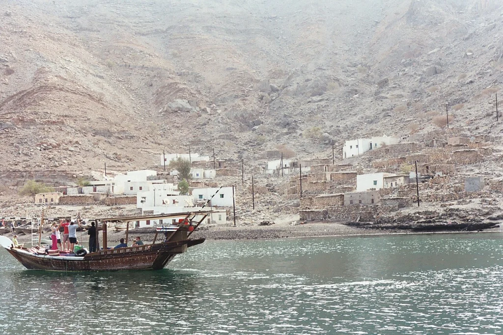 White houses of a fishing village at the foot of bare mountains, with a dhow on the water in front