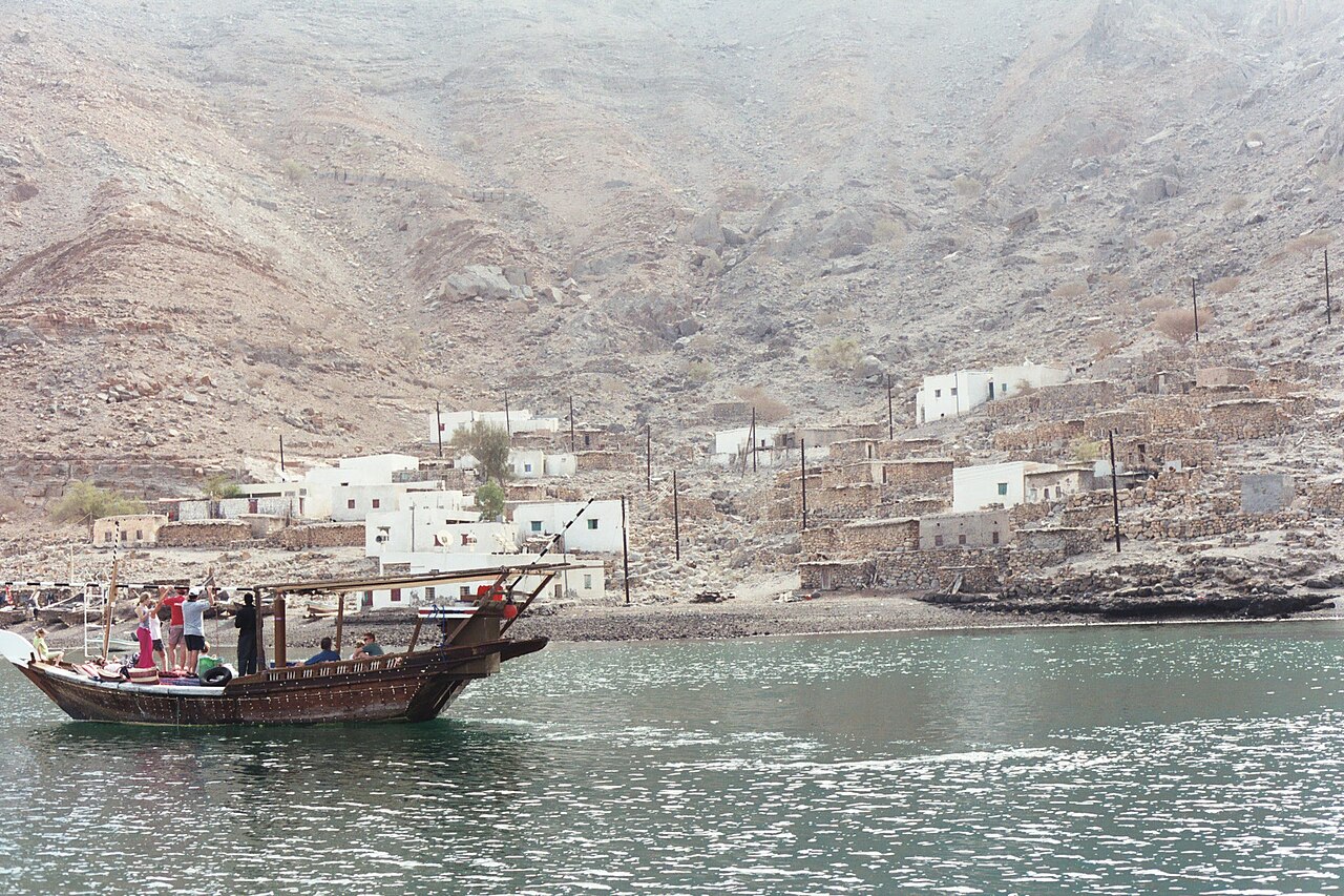 White houses of a fishing village at the foot of bare mountains, with a dhow on the water in front