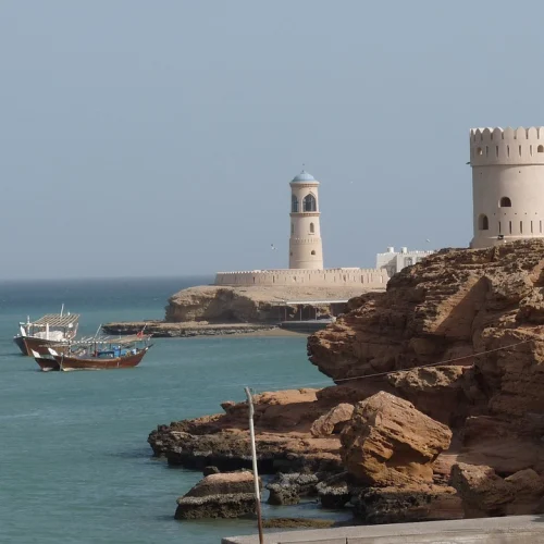 Watchtowers and a lighthouse on a rocky shore, with dhows at anchor